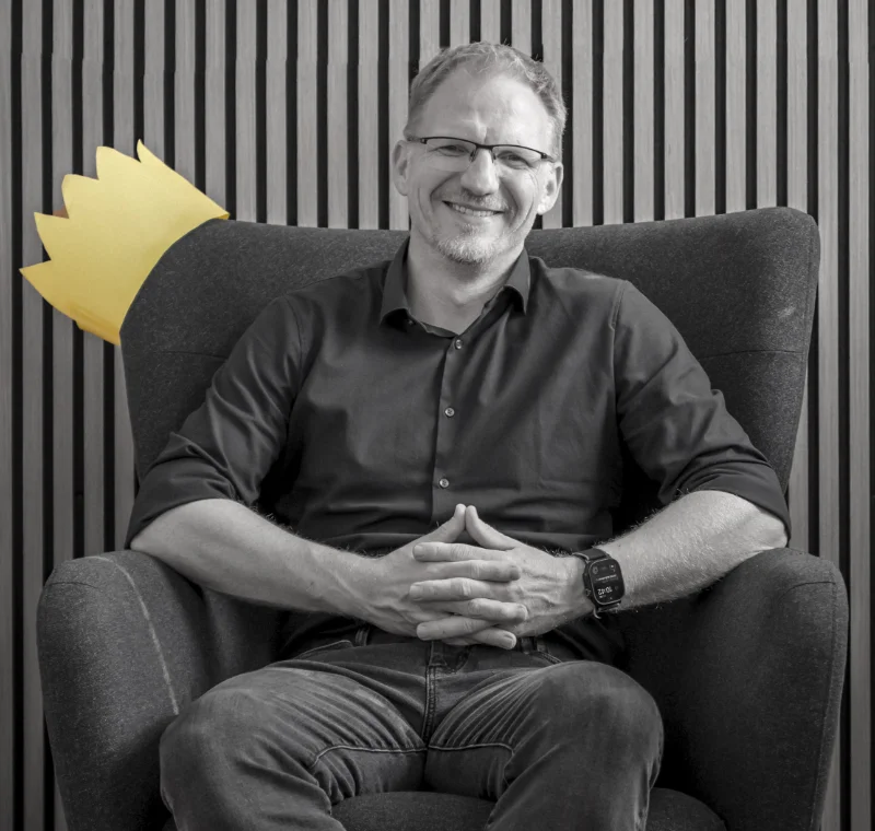 Timo Koenig sitzt entspannt in einem Sessel, hinter ihm eine gelbe Papierkrone